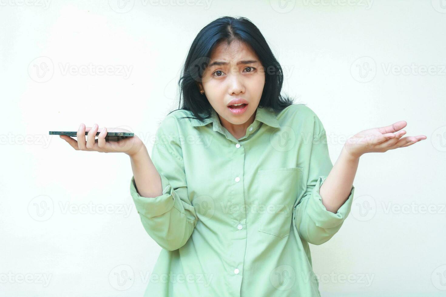 confused asian woman wearing green shirt shrug shoulders and spreads palms holding smartphone looking at camera worried standing over isolated background, considering gesture, I do not know photo