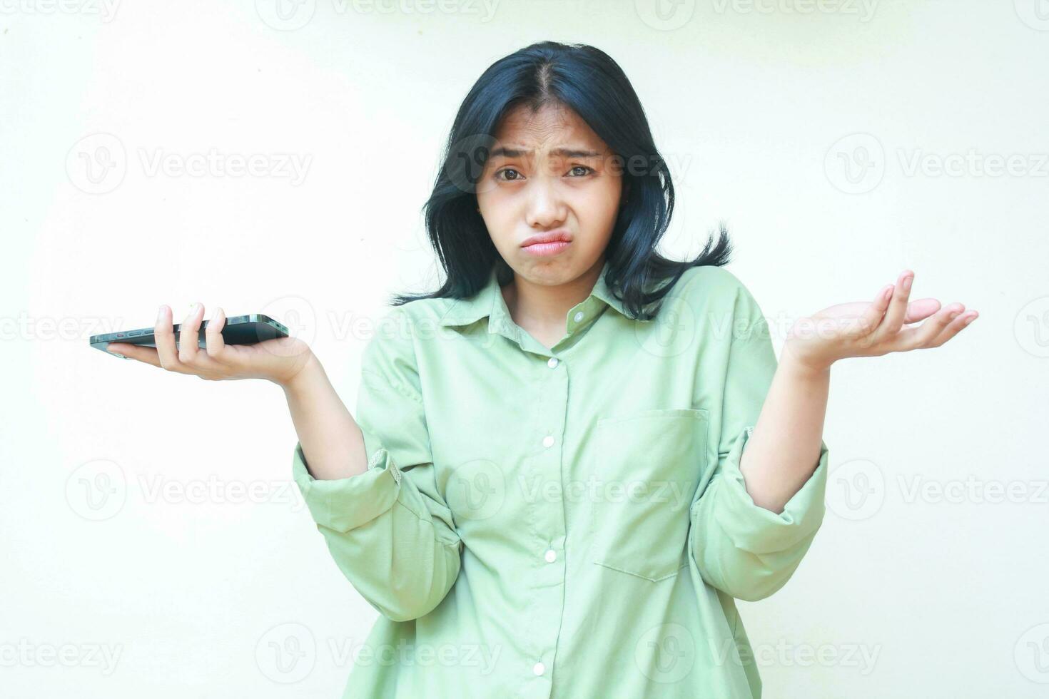 portrait of confused asian woman wearing green shirt shrug shoulders and spreads palms holding smartphone looking at camera worried standing over isolated background, considering gesture photo