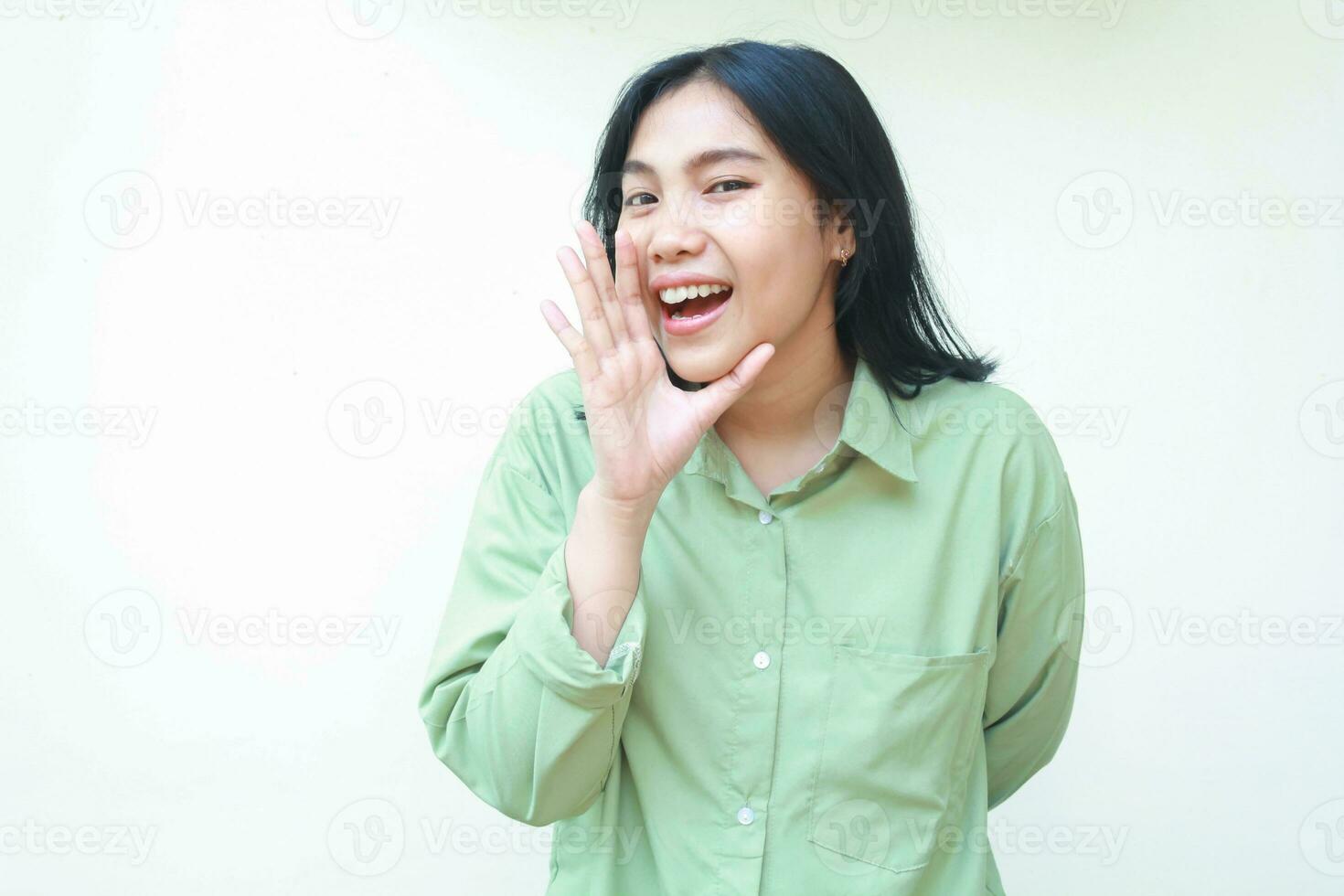 confident pleased asian woman with dark hair raising palms over her open mouth whisperings at camera to announce new product wearing green casual oversized shirt isolated on white background photo