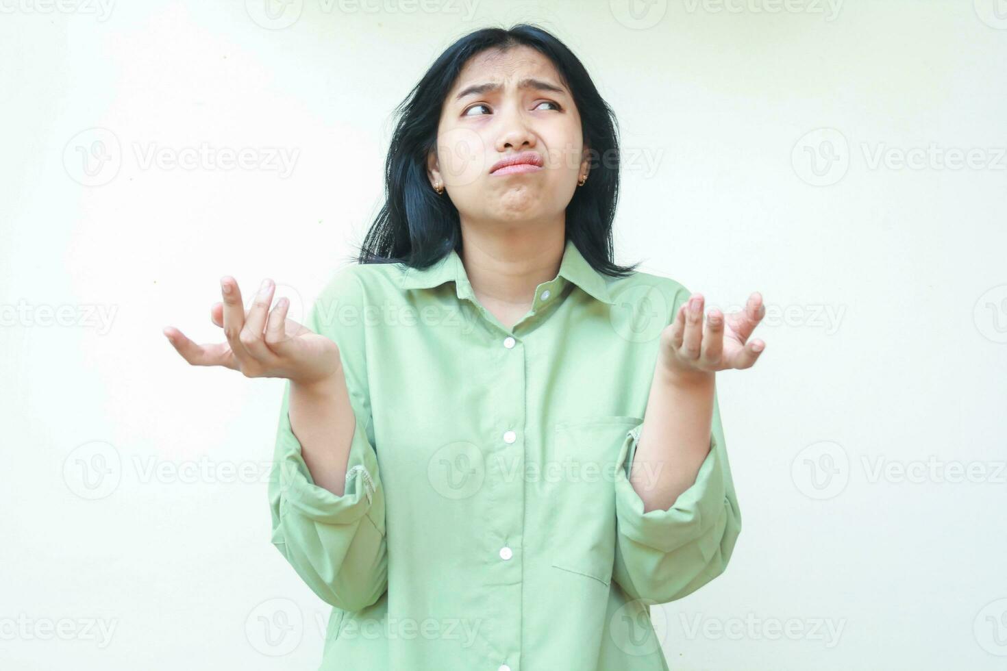 clueless asian woman with dark hair looking at camera with confusion expression raising two palms sideways wearing green oversized shirt, showing and presenting gesture, questioning, asking photo