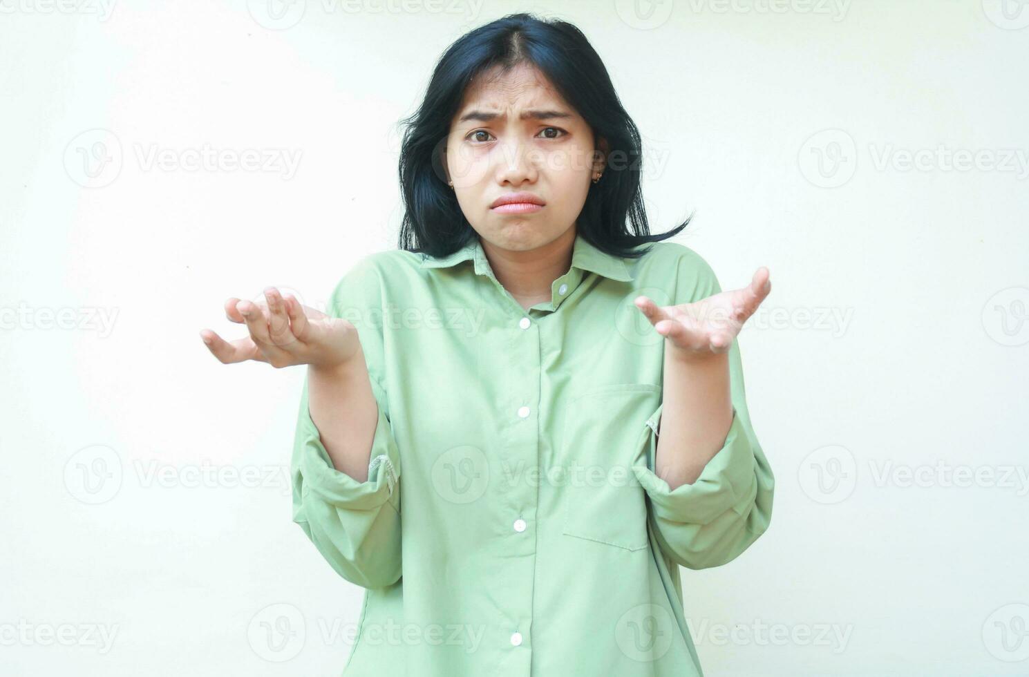 clueless asian woman with dark hair looking at camera with confusion expression raising two palms sideways wearing green oversized shirt, showing and presenting gesture, questioning, asking photo