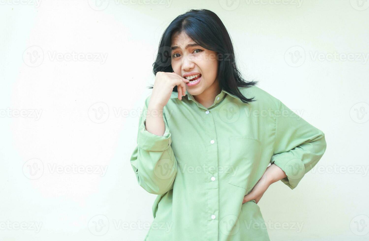asustado asiático mujer mordedura su dedo con manos en cintura mirando a cámara espectáculo asustado cara vistiendo verde de gran tamaño camisa aislado en blanco antecedentes foto