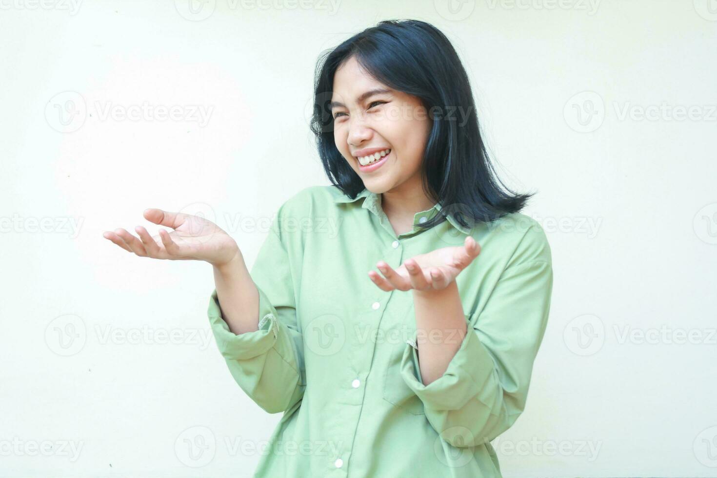 carefree delighted dark hair asian woman raising palms and looking away surprised wearing green oversized shirt isolated on white background, receiving gift gesture photo