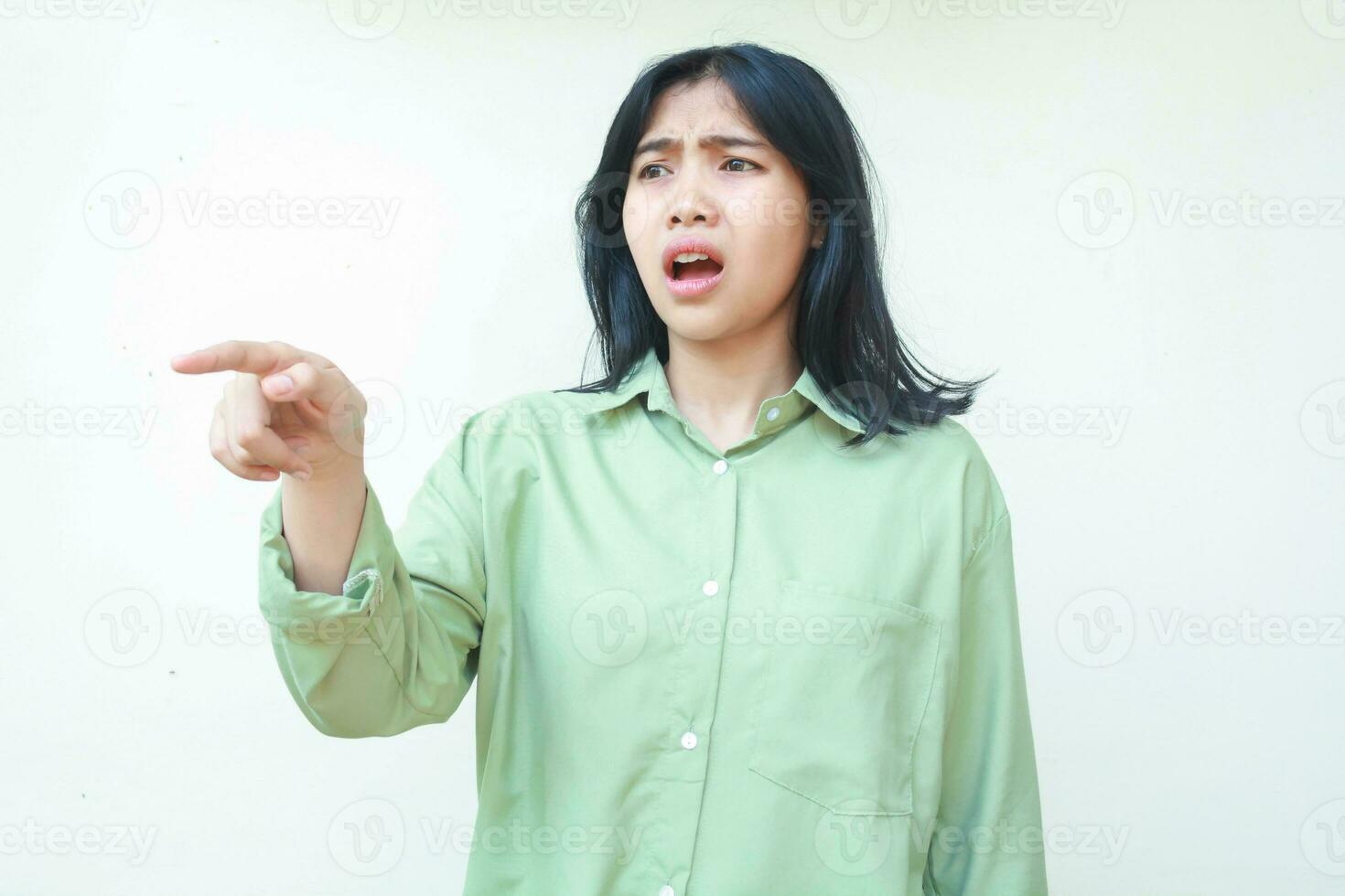 shocked dark hair asian woman pointing finger and looking away stunned with open mouth wearing green oversized shirt isolated on white background photo