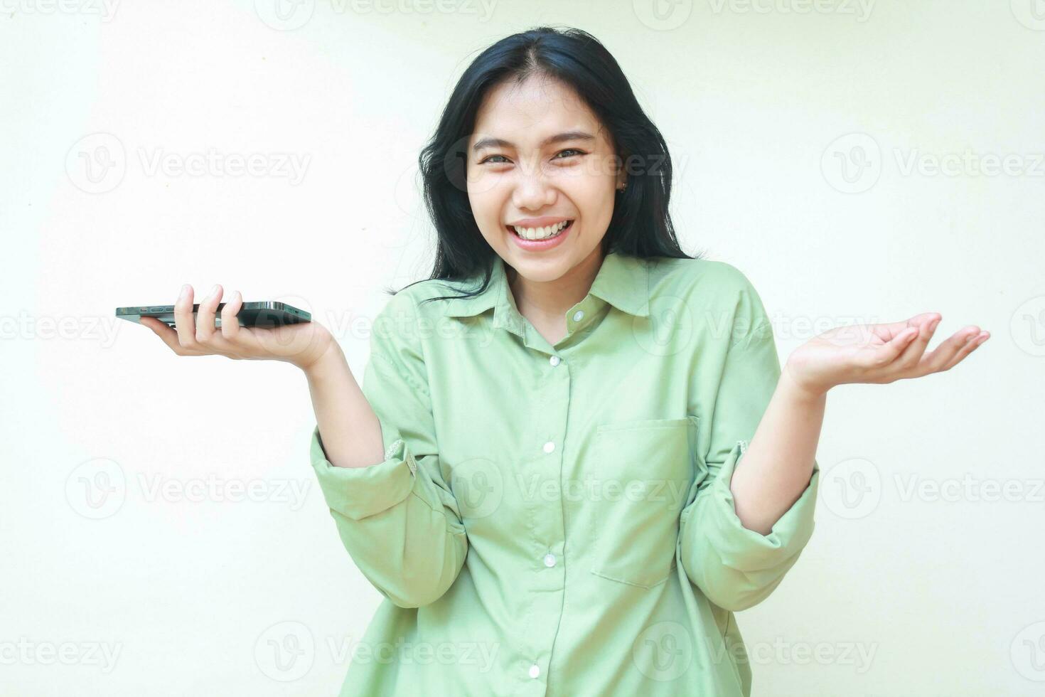 delighted ecstatic pretty asian woman with dark hair wearing green oversize shirt laughing at camera raising hands spreads palms sideways and holding smartphone offering choice isolated photo