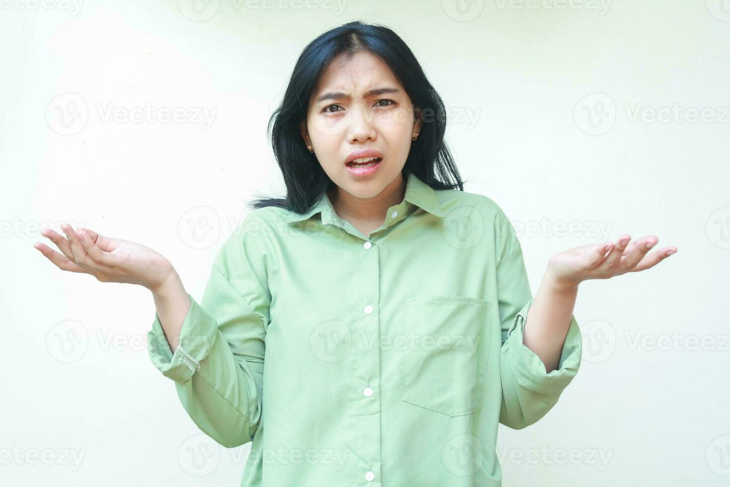 clueless asian woman with dark hair looking at camera with confusion expression raising two palms sideways, showing and presenting gesture, choose product, wearing green shirt isolated on white photo