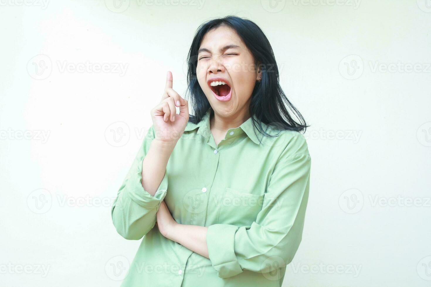 tired yawning asian woman pointing finger up to empty space and folding arms with eyes closed and mouth wide open just wakeup gesture wearing green oversiszed pajamas isolated on white photo