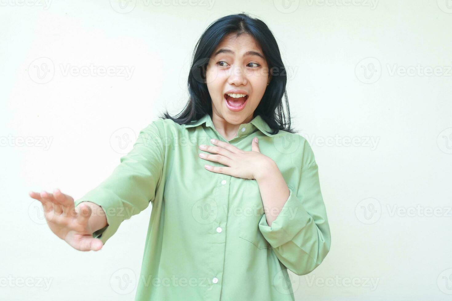 ecstatic carefree asian woman reaching hands forward and touching her chest surprised looking away wearing green oversized shirt isolated on white background photo