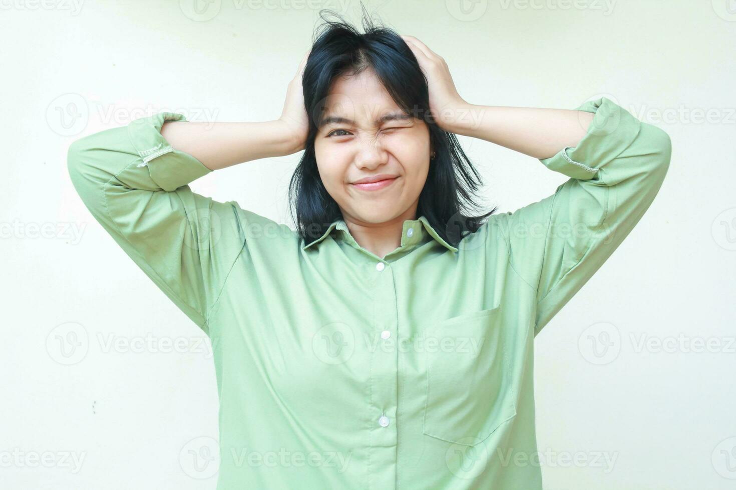 no way. smiling asian woman wearing green over size shirt grabbing her head and closed one eye flrting at camera see something unbeliavable while standing over isolated white background photo