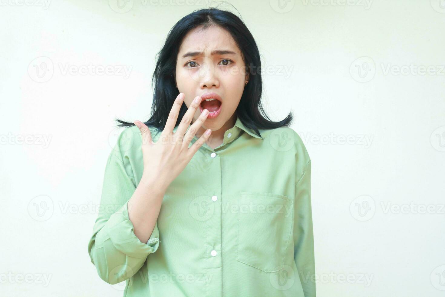 conmocionado oscuro pelo asiático mujer mirando a cámara con abierto boca demostración Guau expresión y cubierta con manos vistiendo verde de gran tamaño camisa aislado en blanco antecedentes foto