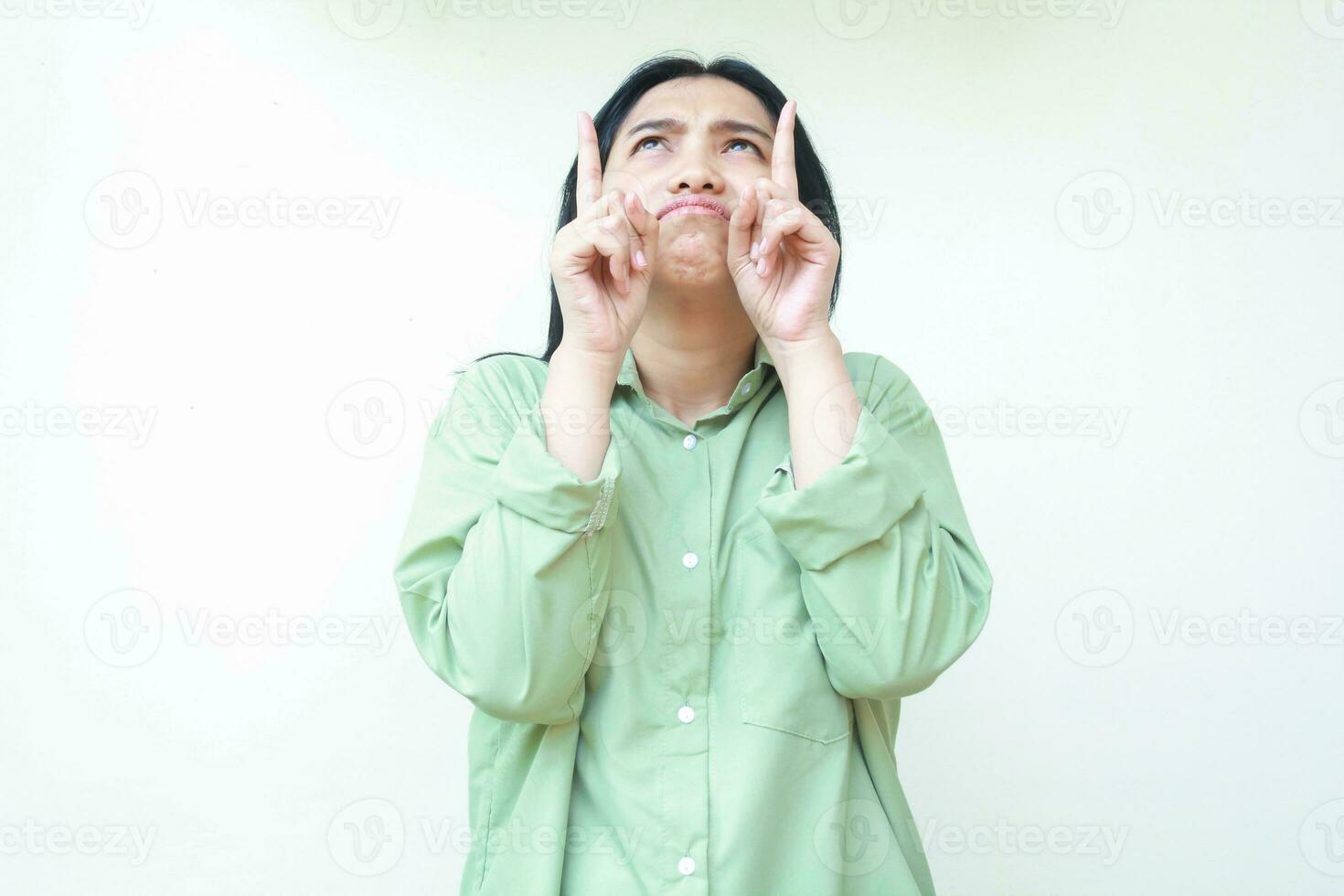 depressed asian woman with dark hair looking above and pointing up to empty space do showing presenting gesture wearing green oversized shirt standing isolated white background photo