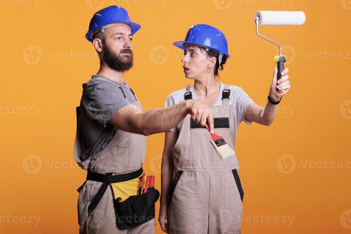 Young construction workers posing with painting tools, standing over yellow background. Team of constructors holding paintbrush and roller brush to work on refurbishment project in studio. photo