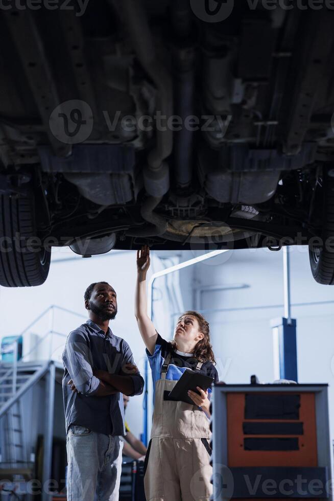 Engineer in car service uses tablet to calculate invoice after fixing broken automobile tires. Worker uses device to inform client of final costs after mending vehicle suspended on overhead lift photo