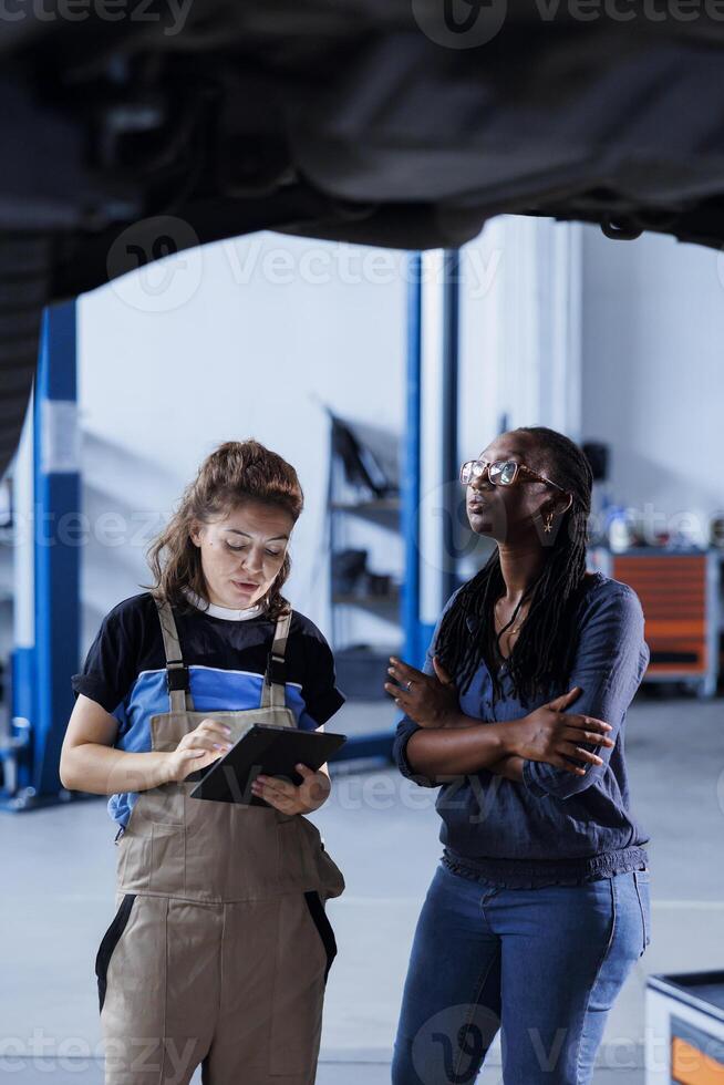 Repairman in car service orders new tires for damaged vehicle using tablet. Engineer next to client looking online for components to replace old ones in malfunctioning suspended automobile photo