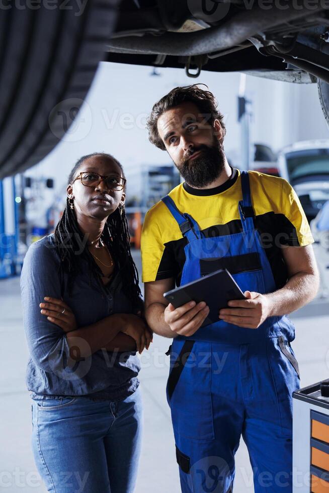 Proficient professional helping customer with car checkup in auto repair shop. Garage employee looking over automobile parts with woman, repairing her busted vehicle wheels during inspection photo