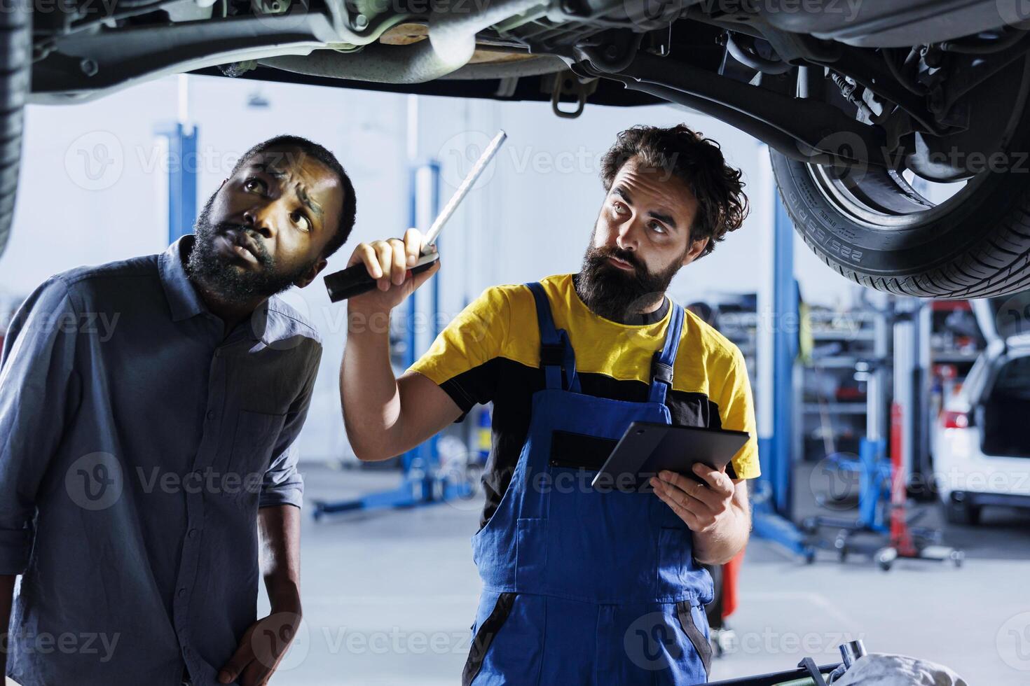 Repairman in car service uses tablet to calculate invoice after fixing broken automobile wheels. Professional uses device to inform client of final costs after mending vehicle suspended on overhead photo