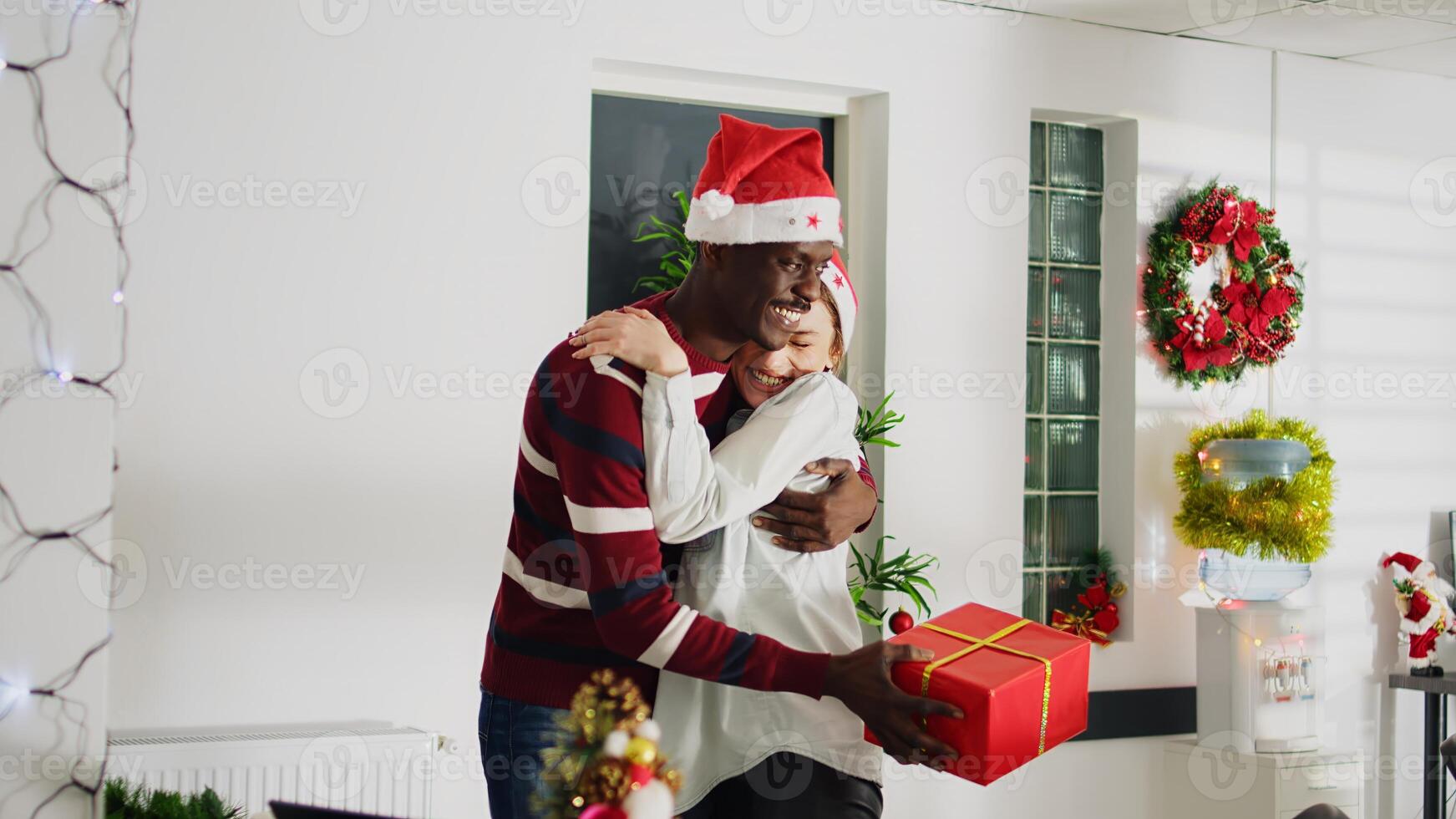 Dolly in shot of employee surprised by coworker with Christmas gift, hugging eachother in festive decorated office. Staff member excited after being offered xmas present at work, shaking box photo