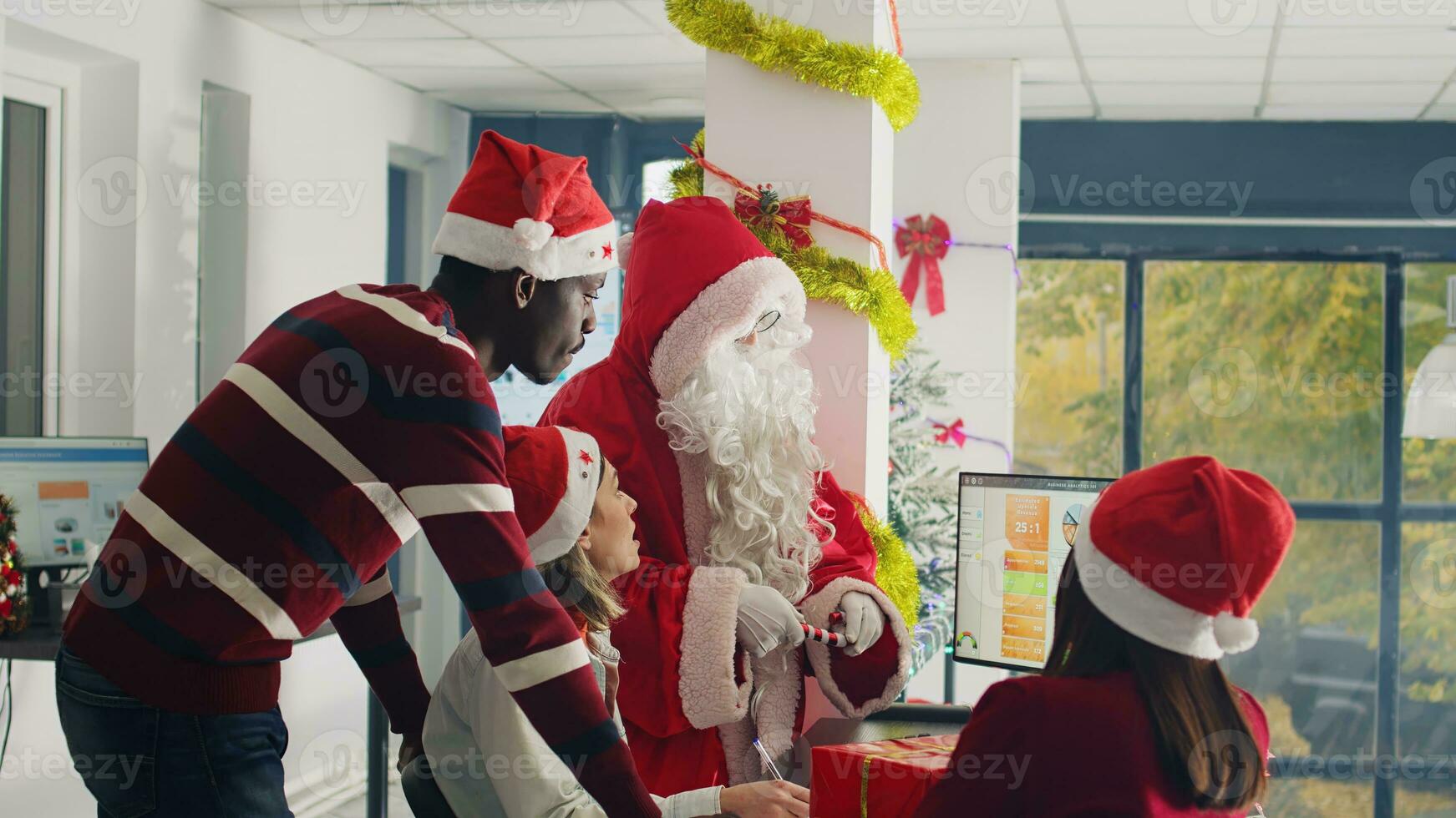 Manager dressed as Santa Claus in xmas ornate office using candy cane ornament as pointer while showing numbers on screen. Supervisor wearing Christmas costume overseeing work done by his team photo