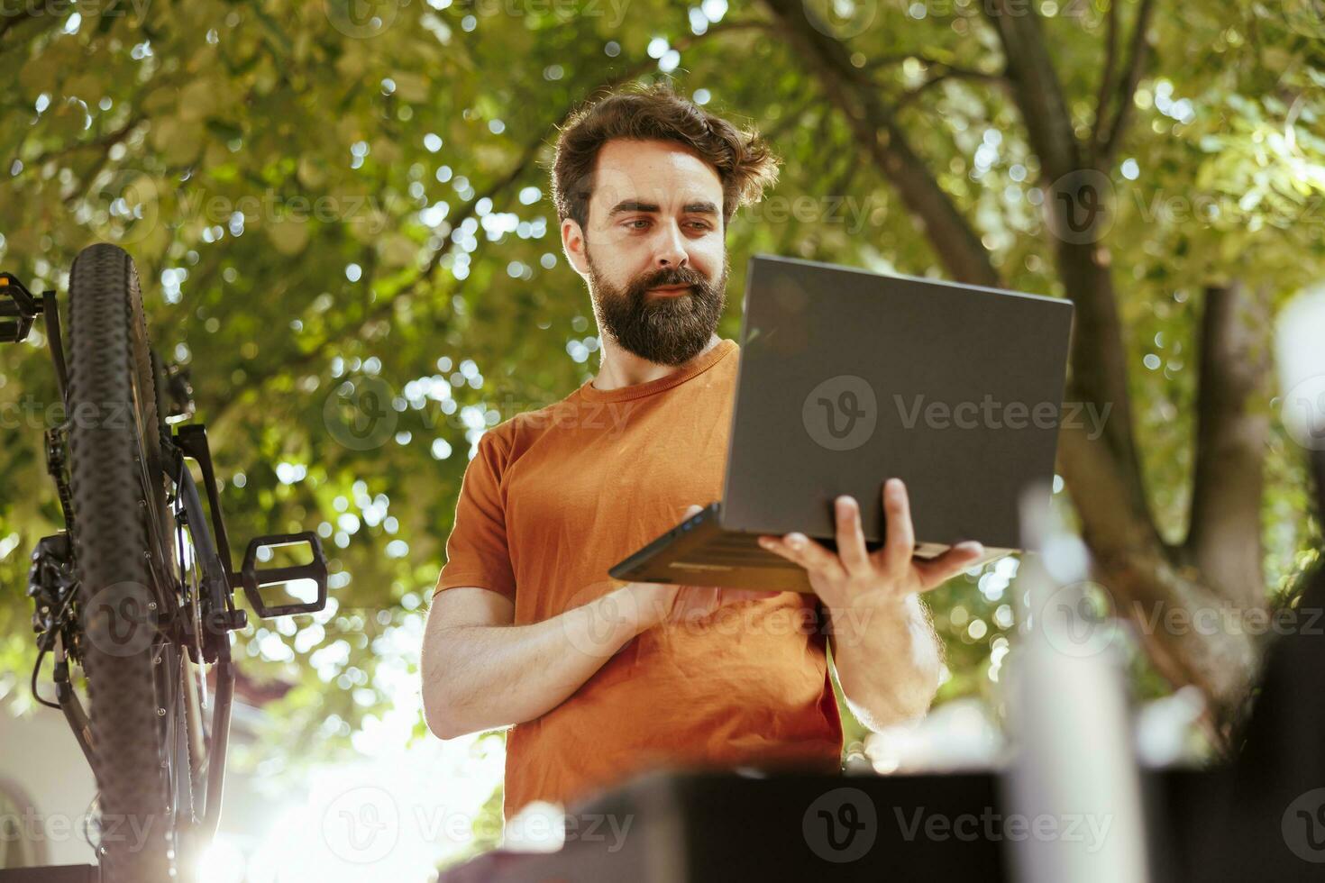 Healthy dedicated caucasian man searching on laptop to fix broken modern bicycle. Active young male cyclist ensures bike components are secure for summer outdoor leisure cycling. photo