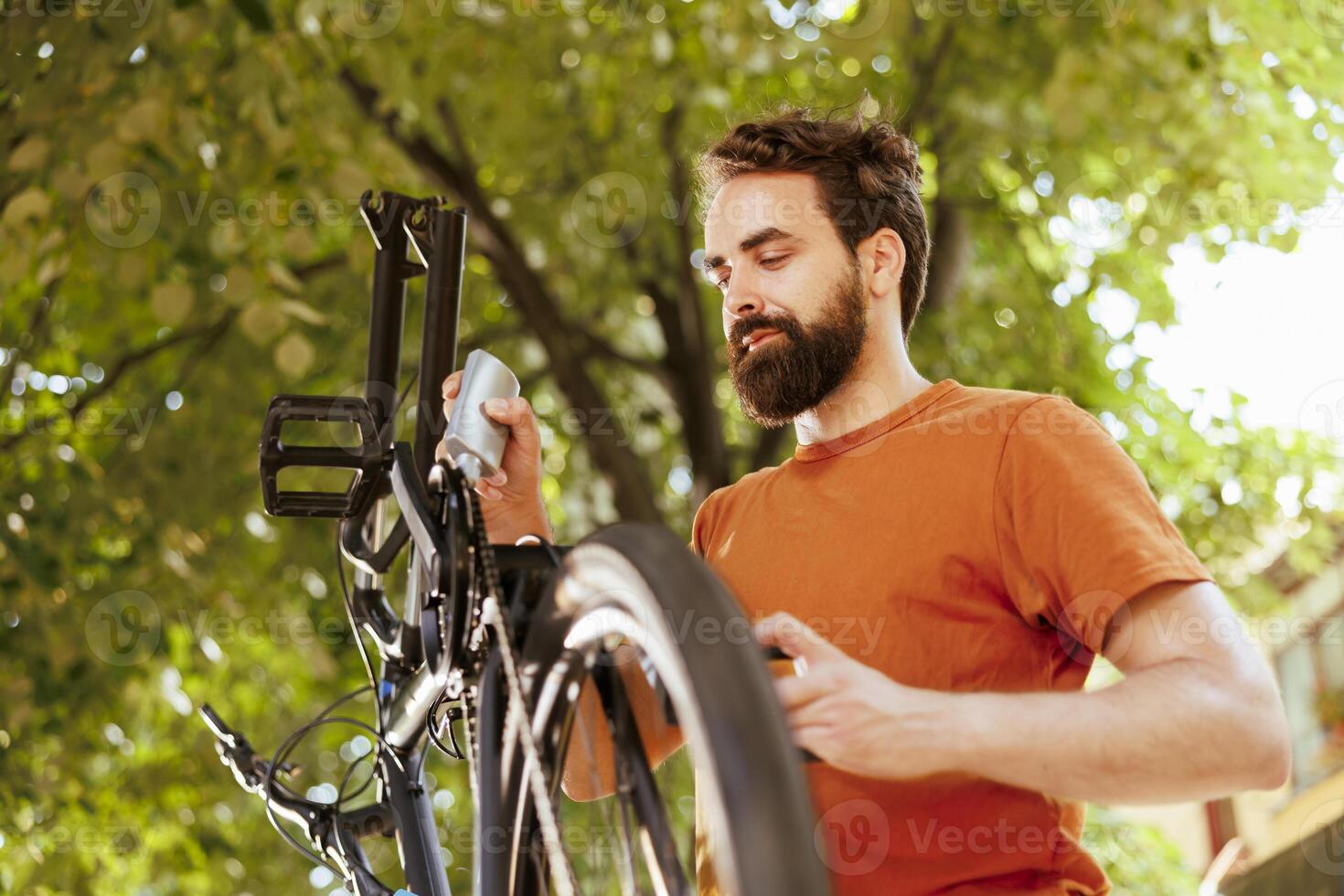 activo y sano masculino ciclista inspeccionando su bicicletas rueda para daños y perjuicios en el patio trasero. imagen retrata joven caucásico hombre utilizando lubricante a reparar roto componente asegurando un seguro y seguro paseo foto