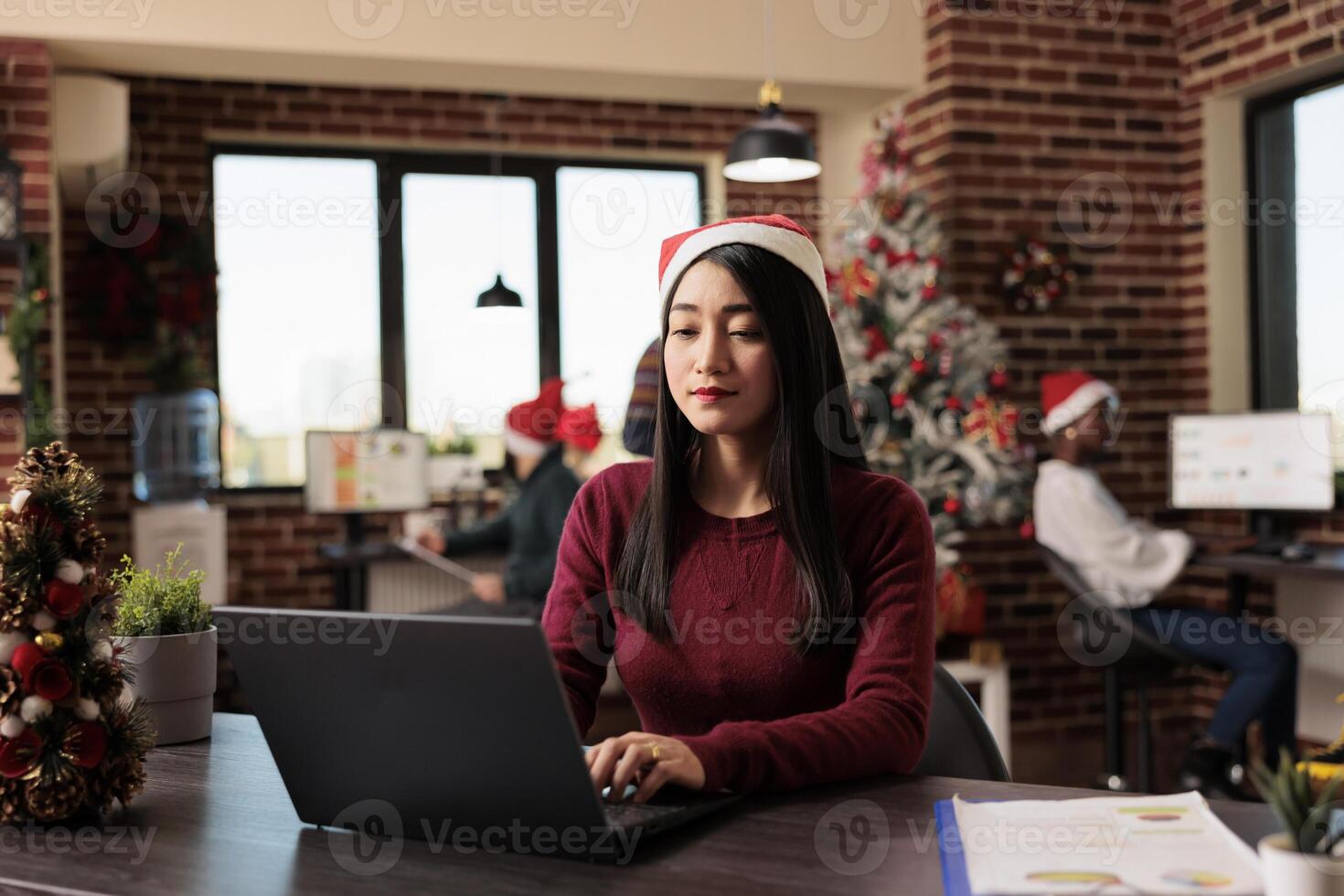 Asian woman in santa hat managing project deadline on laptop while working in decorated office at christmas holdiay. Company employee using computer at workplace during winter celebration season photo