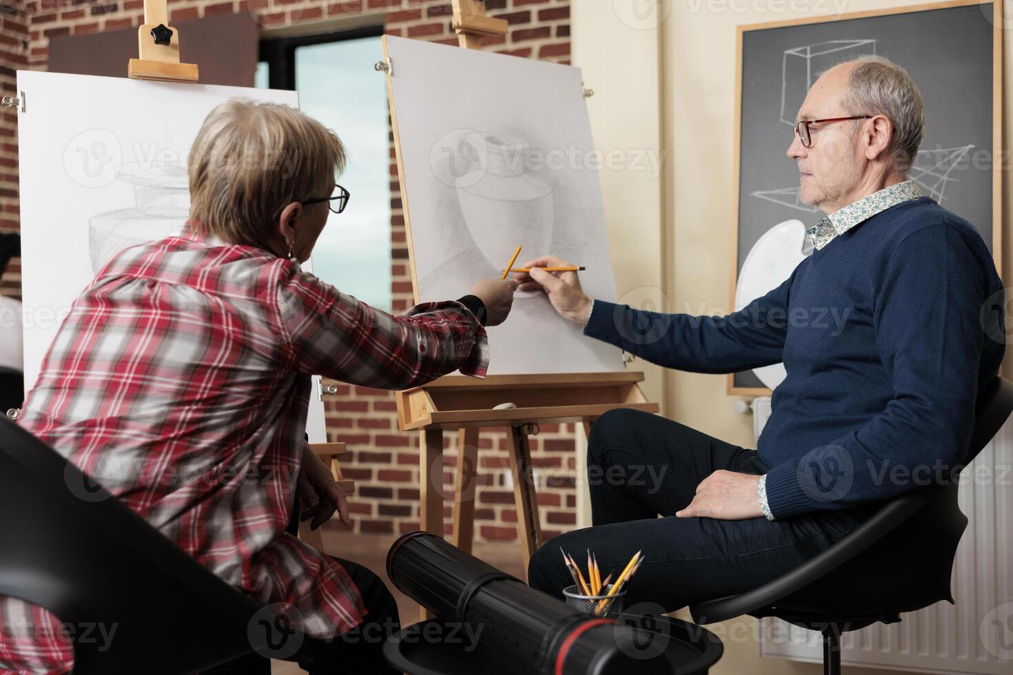 Retired couple talking discussing artwork while learning new skill. Senior man and woman sitting at easels learn to draw, attending group art class together. Creative activities to do with family photo