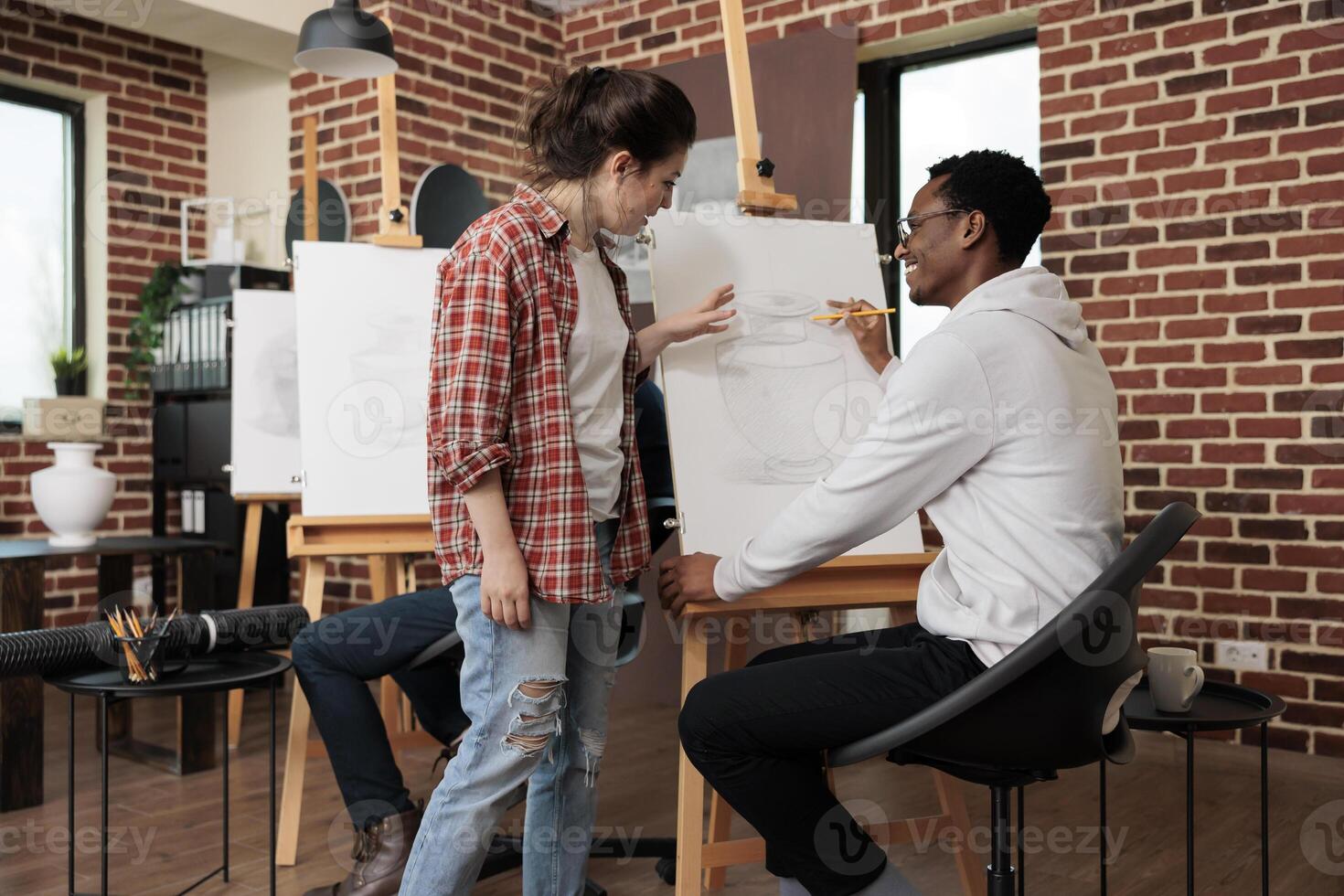 Young positive African American guy making friends and new connections at group art class, talking with woman teacher while drawing with pencil on canvas. Learning new skills and meeting people photo