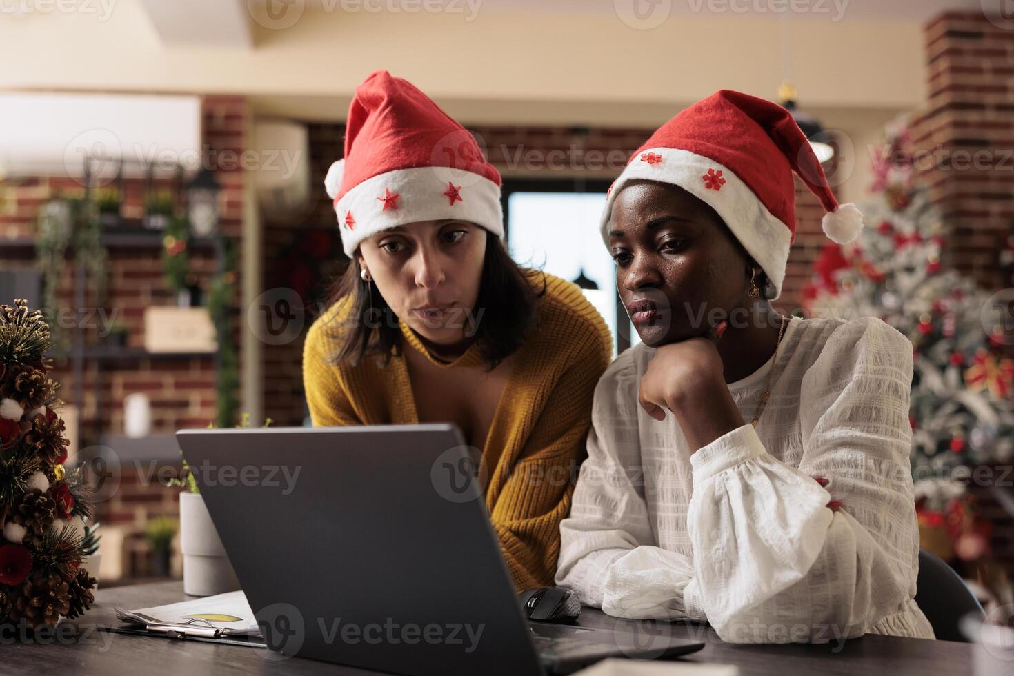 Two women coworkers checking project report on laptop and celebrating xmas in decorated office. African american and caucasian employees in santa hats working on startup task together photo