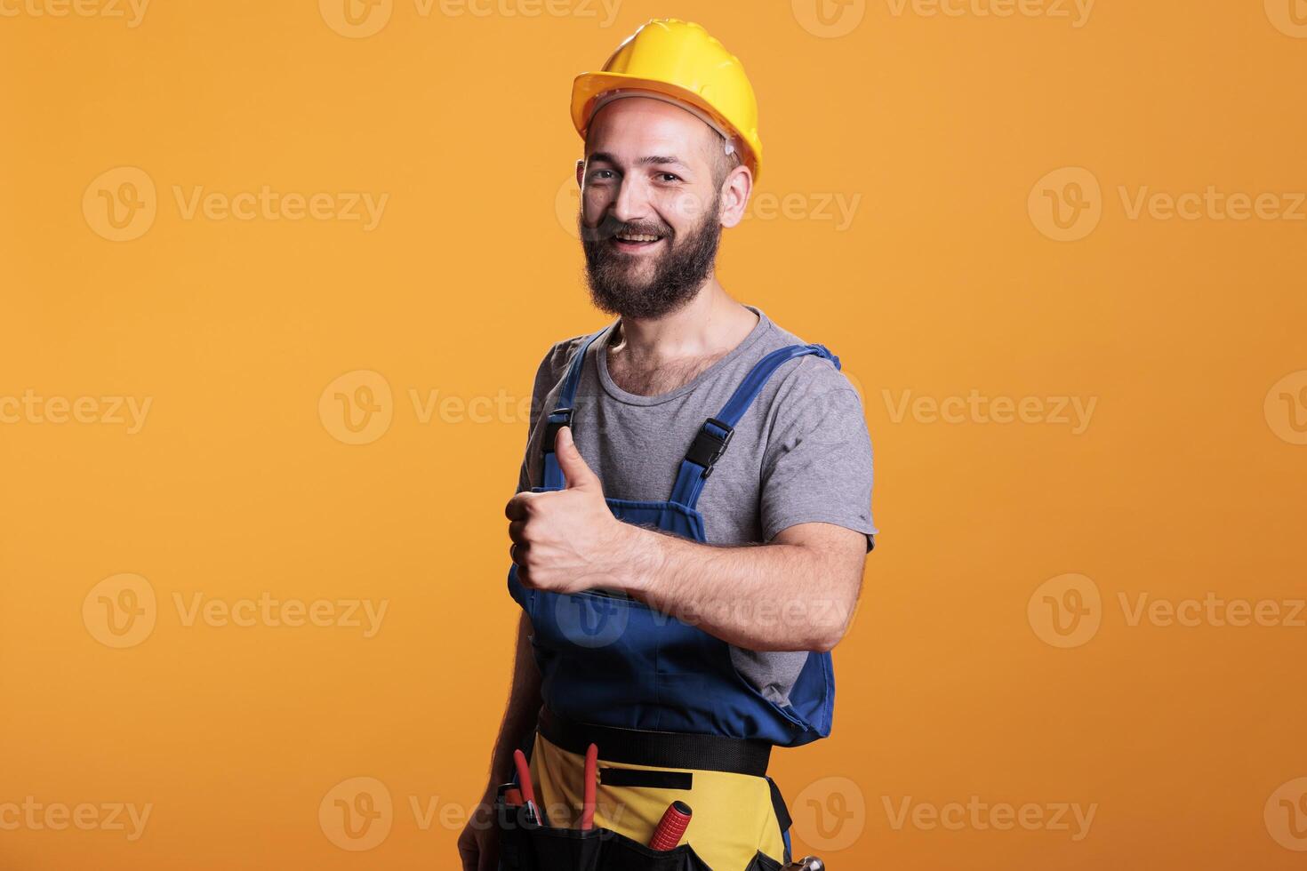 Young building contractor giving thumbs up on camera, showing like and approval gesture in studio. Male construction worker expressing agreement and good symbol, confident happy expert. photo