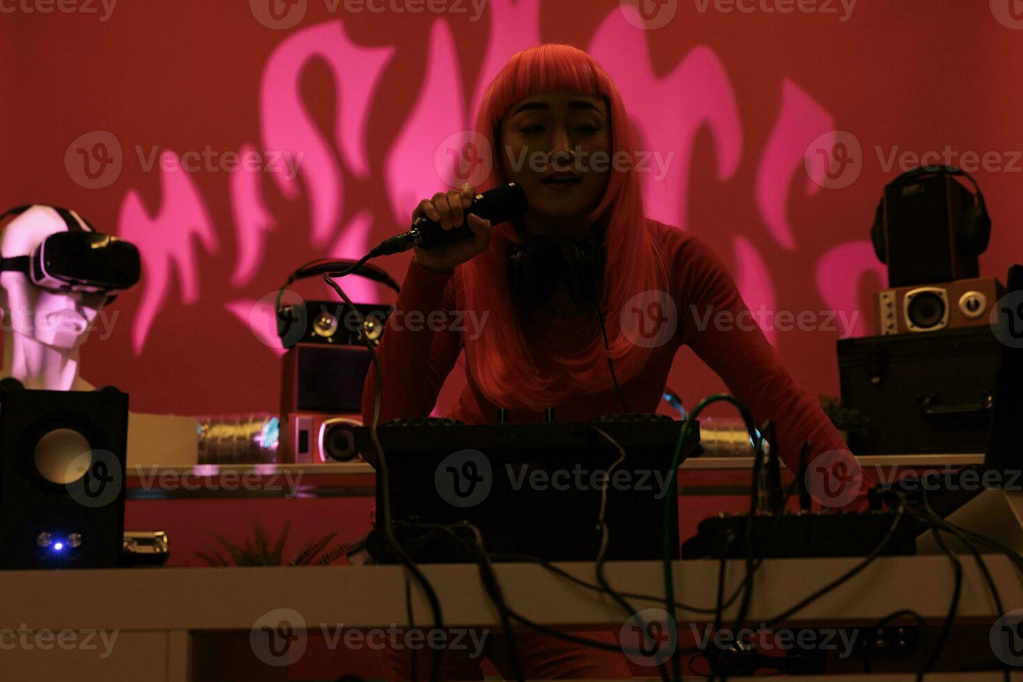 Cheeful artist with pink hair standing at dj table playing electronic music with turntables, enjoying to perform at night in club. Asian performer having fun with fans during techno concert photo