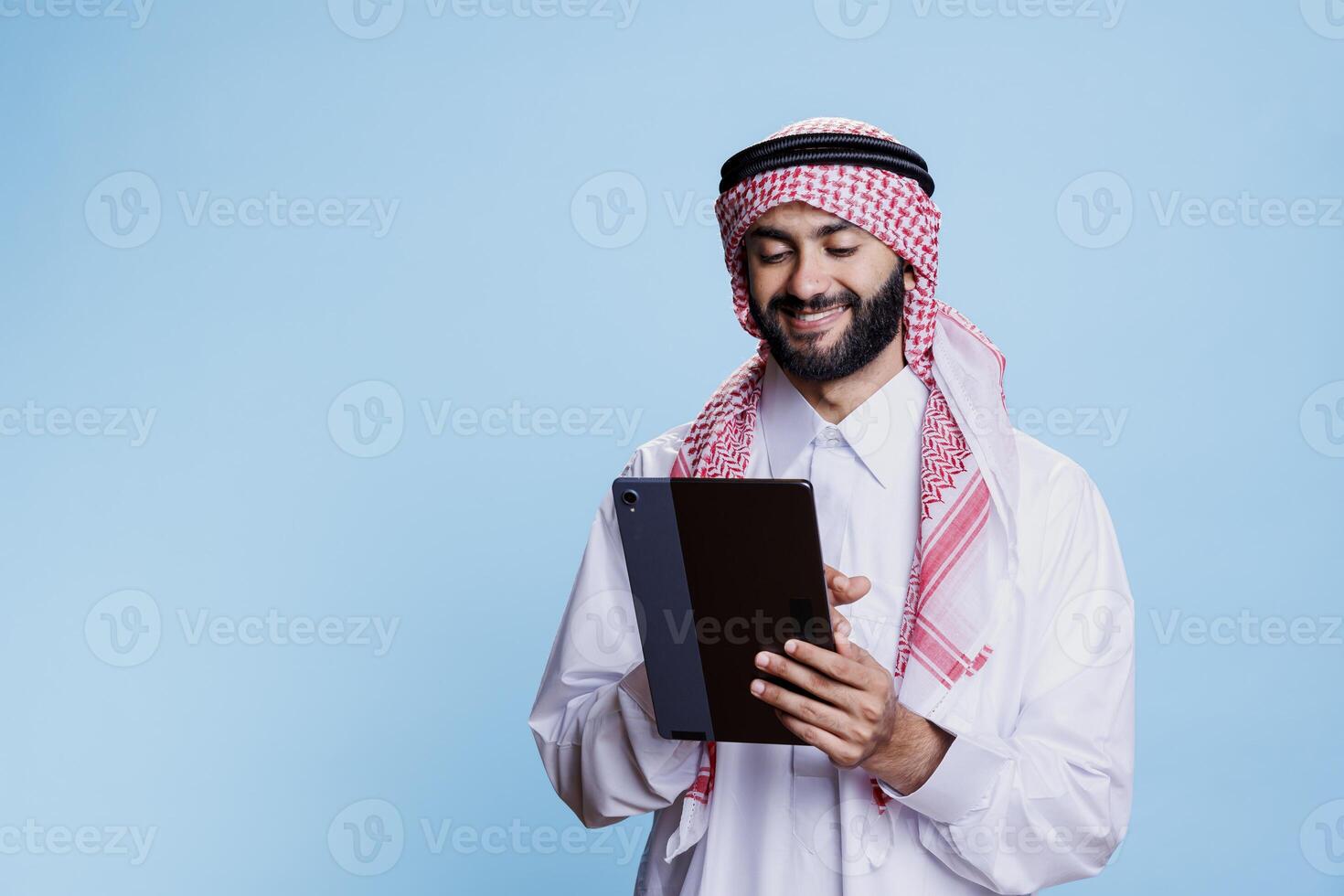 Smiling man wearing muslim headdress and thobe holding digital tablet and tapping on touchscreen. Arab person scrolling internet page on portable gadget with cheerful expression photo