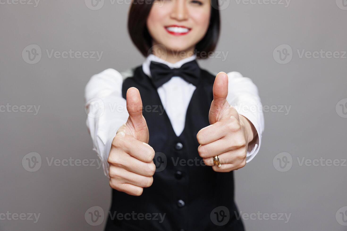 Cheerful waitress showing thumbs up gesture with closeup on arms. Young asian woman receptionist sharing restaurant service positive experience with approval symbol in studio photo