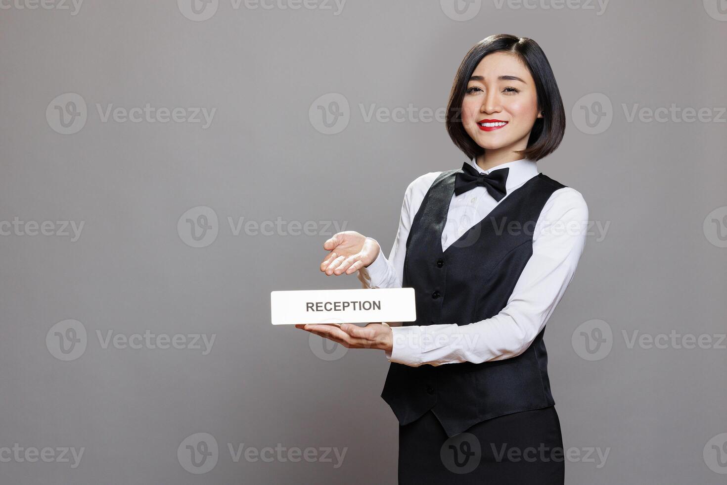 Hotel smiling woman employee showing reception plate studio portrait. Friendly asian receptionist wearing professional uniform pointing at steel name signage and looking at camera photo