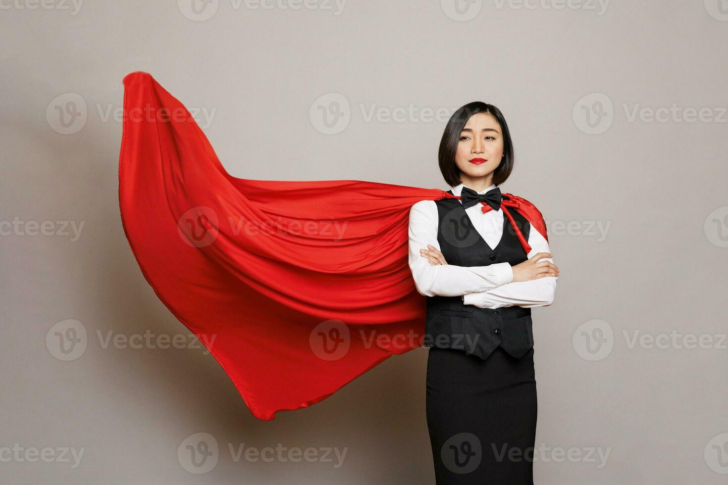 Confident asian waitress standing with folded arms and posing in fluttering superman red cape. Young attractive woman receptionist dressed in hero cloak and catering service uniform photo