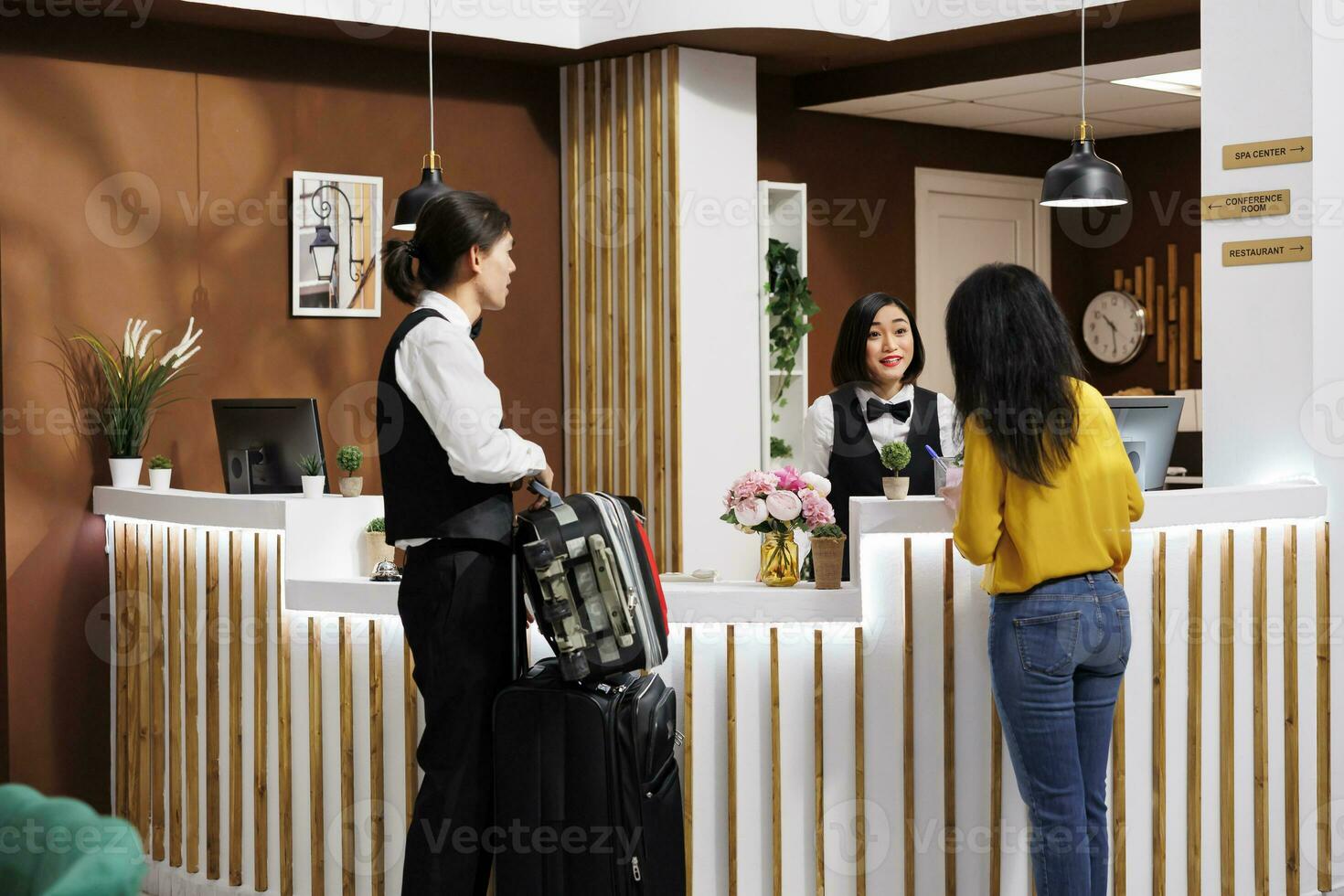 Friendly hotel staff helps client settle in, offering professional support and preparing to take her baggage to room. Asian concierge employee and customer having pleasant talk in foyer photo