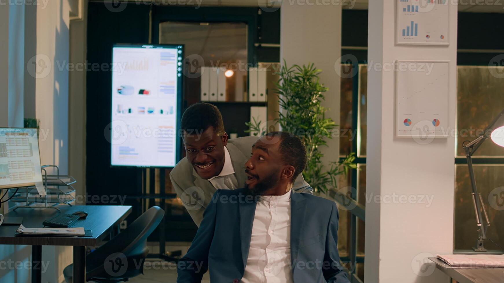 Businessman playing prank on unsuspecting coworker at work, pushing his desk chair. Relaxed staff personnel having fun during nightshift in office, taking break from work photo