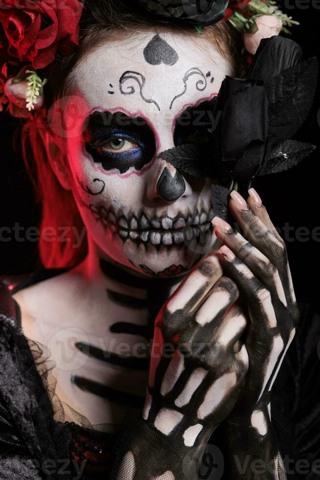 Beautiful lady of dead holding black roses in studio, looking like la cavalera catrina skull with make up. Santa muerte goddess with traditional halloween costume on dios de los muertos. photo