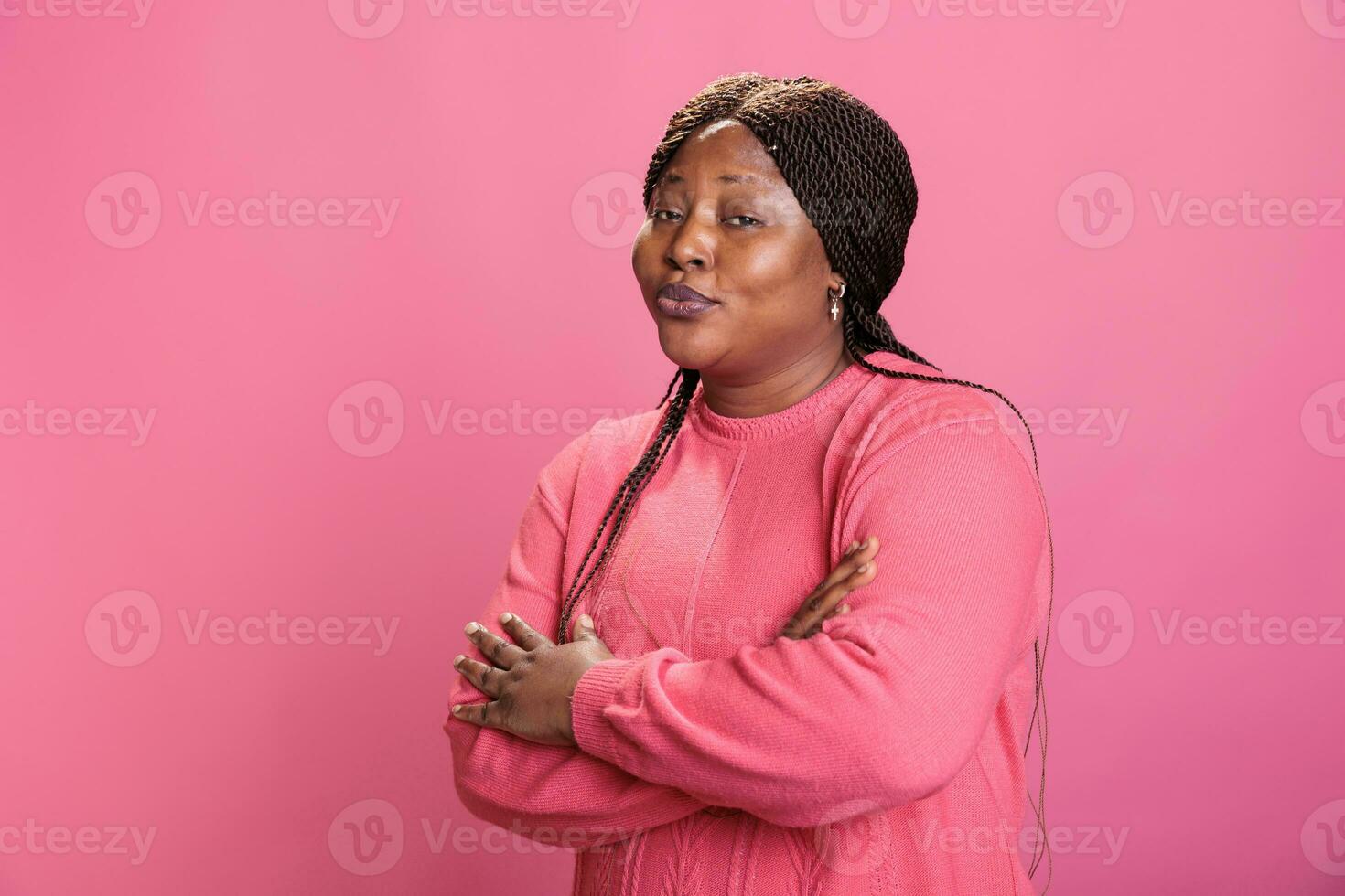 Portrait of excited friendly young adult standing with arm crossed smiling at camera during shoot time posing with confidence in studio with pink background. Cheerful woman with charming expression photo