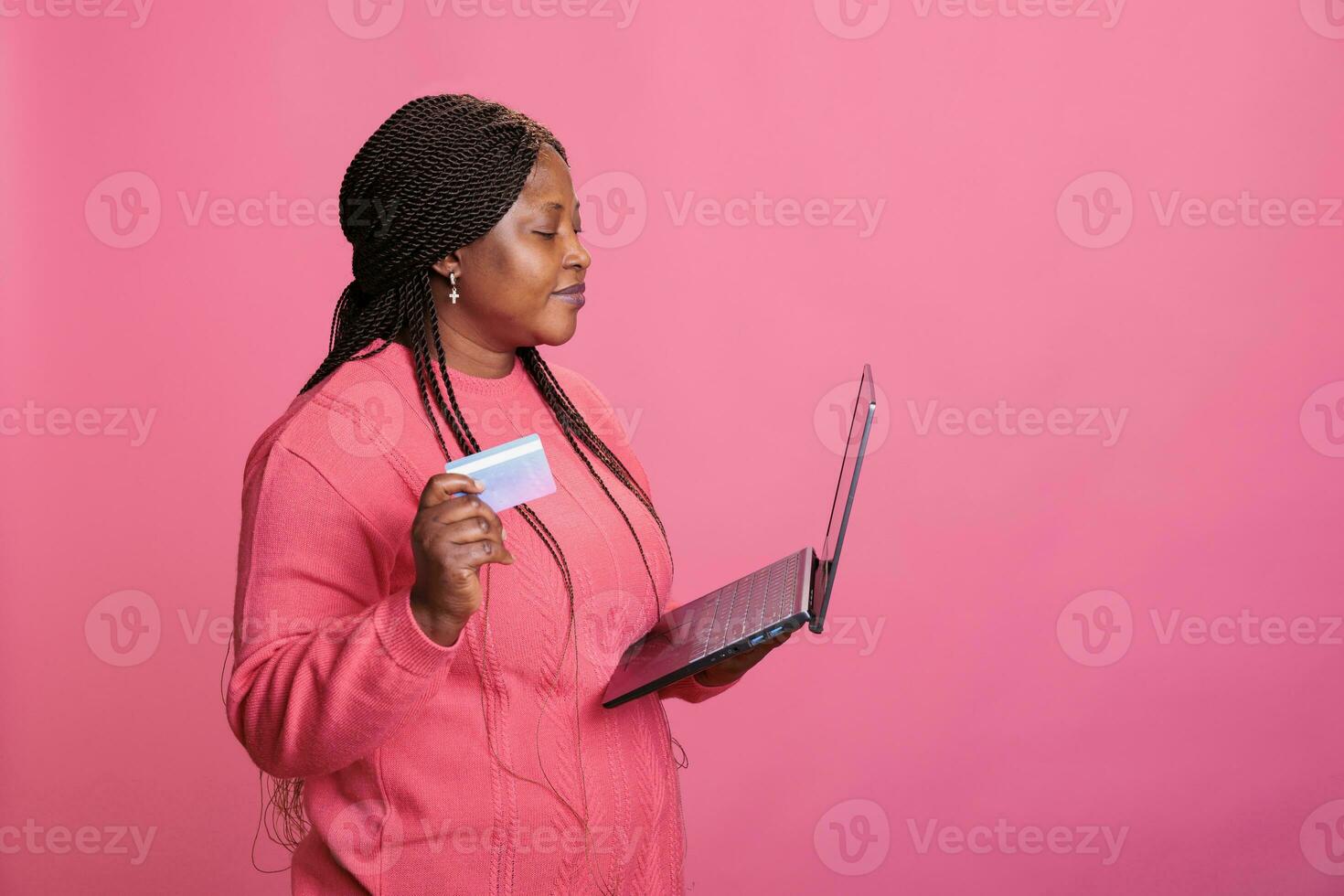 Excited woman inserting credit card information on laptop computer after purchasing fashion items standing in studio over isolated background. Model doing online shopping paying for products photo