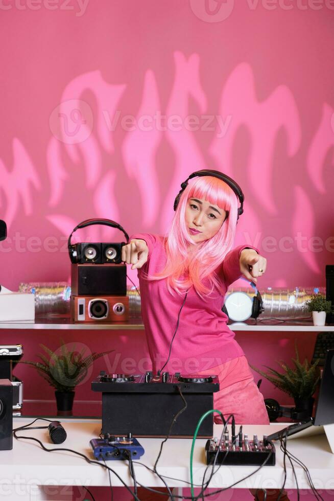 Musical artist standing at dj table mixing techno song with electronic using mixer console, performing music in front of crowd. Asian aerformer with pink hair enjoying to perform at night in club photo