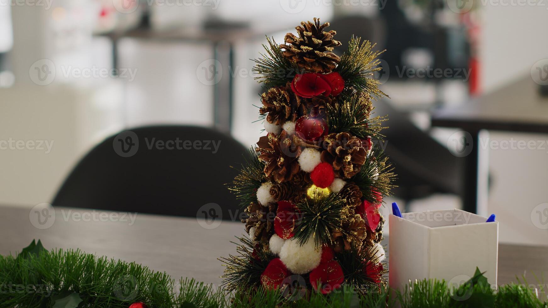 cerca arriba Disparo en miniatura Navidad pino árbol decoración metido en vacío puesta en marcha oficina, con borroso antecedentes de festivo lugar de trabajo lleno con Navidad luces, guirnaldas y coronas foto