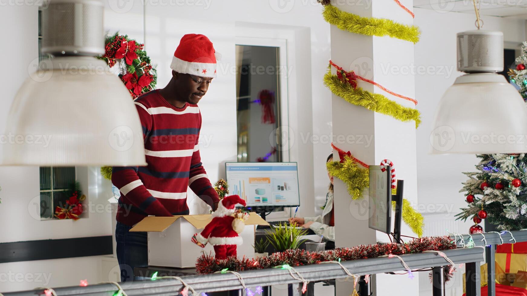 Disappointed worker in festive decorated office packs desk belongings on last day at work, consoled by coworker. Man getting fired from job during Christmas year end layoffs, clearing workstation photo