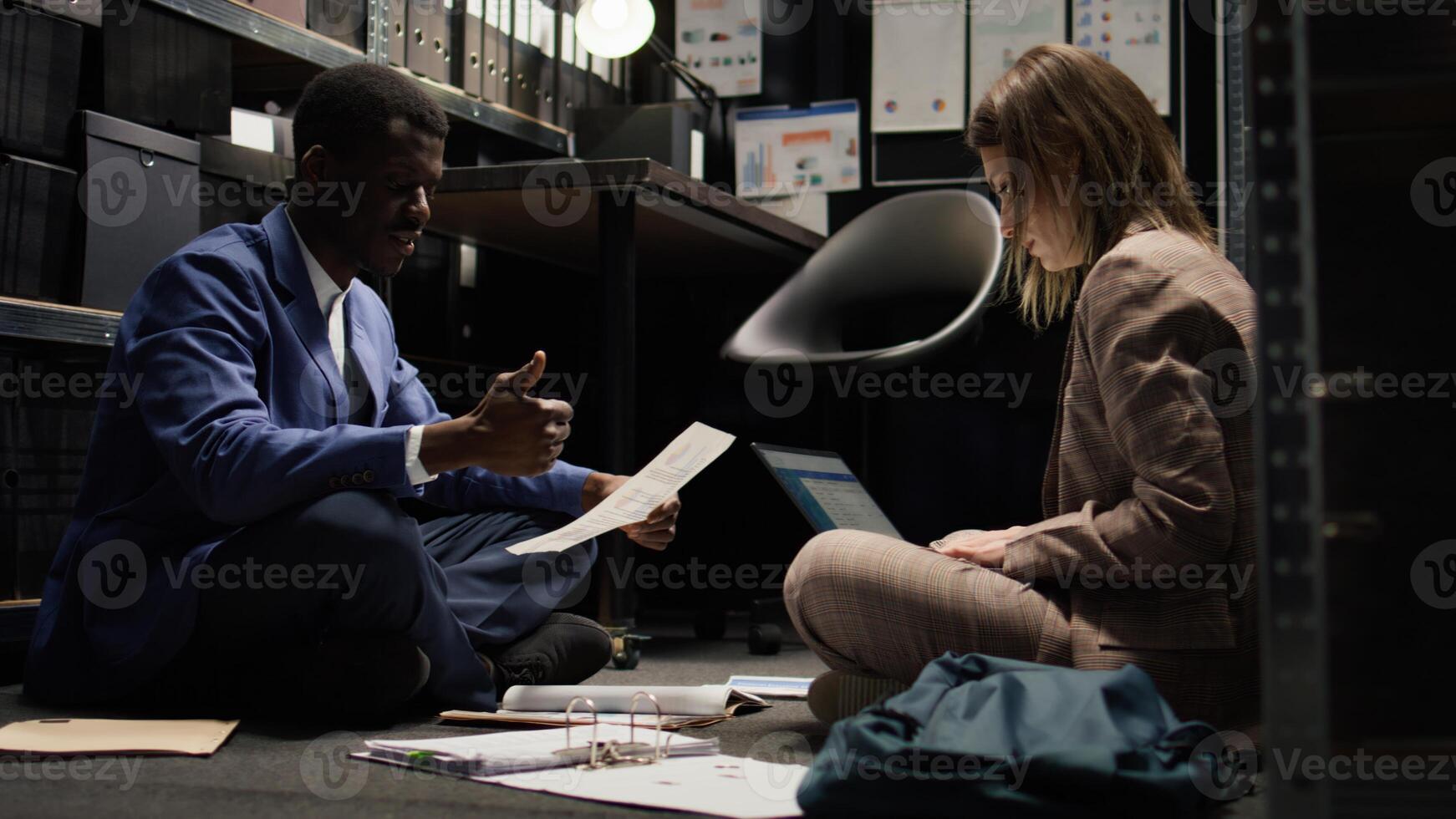 Male inspector and female police officer seated on office floor with paperwork and laptop, conducting crime investigation. Case clues and evidence are examined and discussed by multiracial detectives. photo