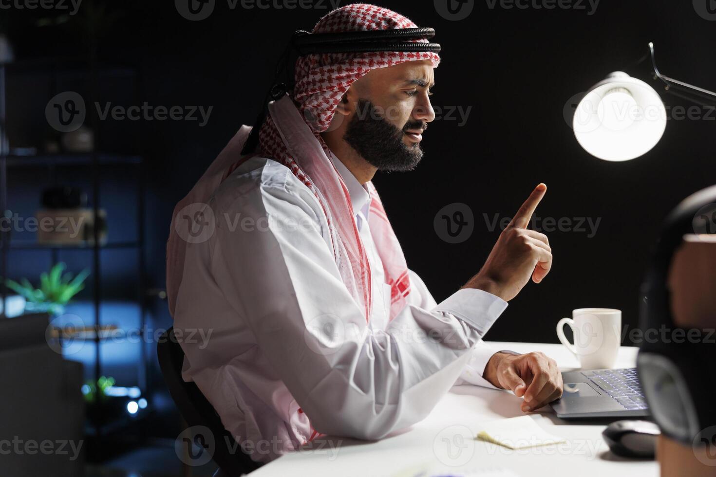 vista lateral Disparo de islámico chico vistiendo tradicional ropa sentado a su escritorio teniendo un conferencia llamada en su ordenador portátil. imagen muestra árabe persona de libre dedicación absorto en un conversacion en su personal computadora. foto