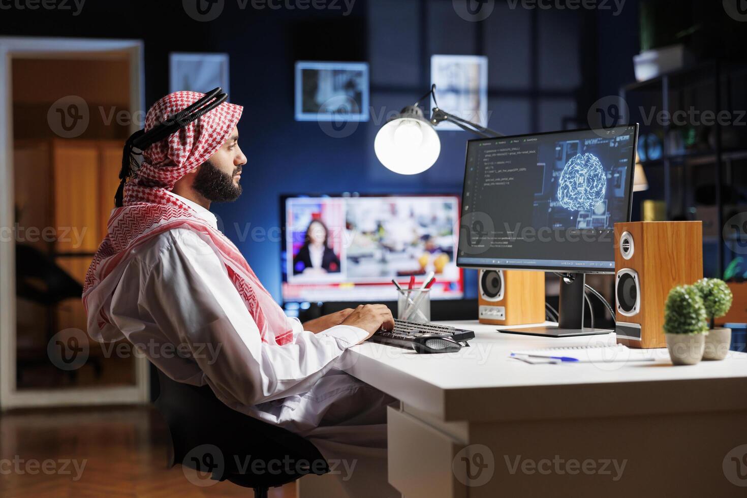 A professional man dressed in Arabic clothing, is working on futuristic interface while seated at his home office desk. Muslim guy manages data, monitors security, and programs AI on desktop computer. photo