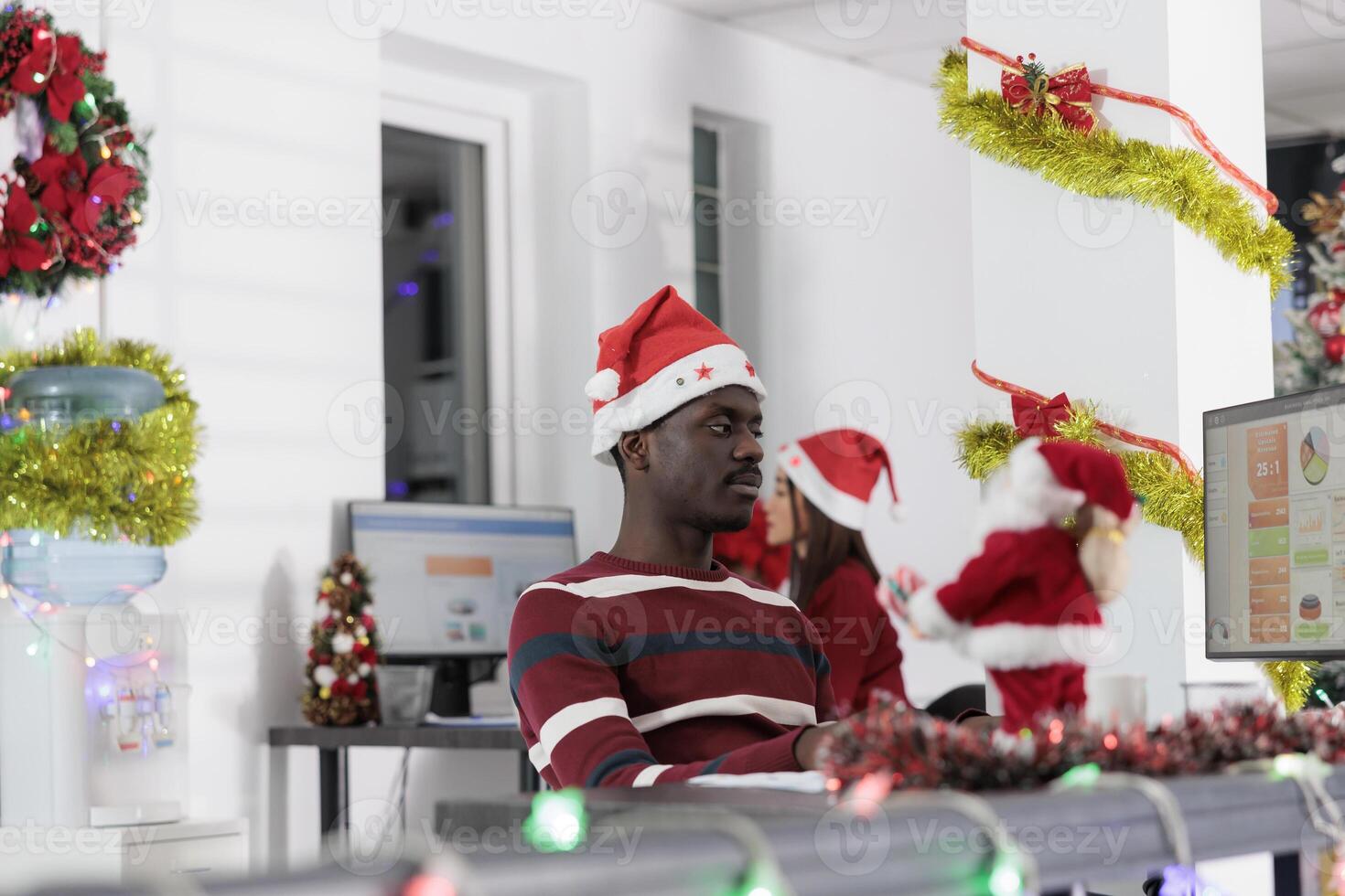 Tired african american employee working on stressful job deadline before Christmas. Workaholic businessman solving project tasks sitting at desk in festive decorated office photo