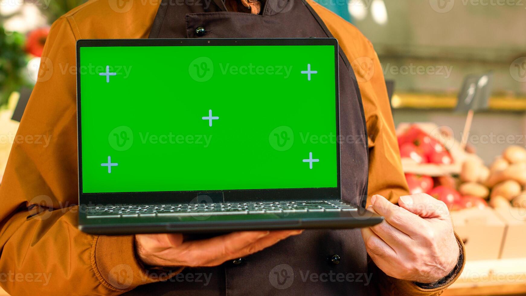 Senior seller showing greenscreen display on laptop, wearing apron at farmers market shop. Male vendor hoding pc with isoated chroma key and blank copyspace mockup template. Close up. photo