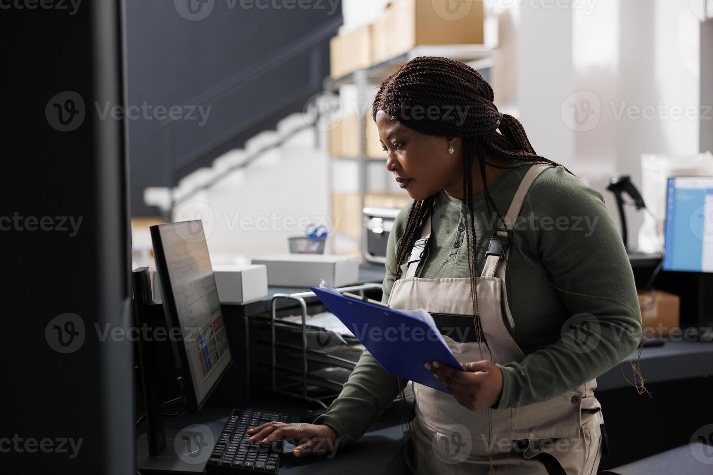 Stockroom supervisor looking at inventory report on computer, working at merchandise quality control in warehouse. African american employee preparing customers orders for shipping in storehouse photo