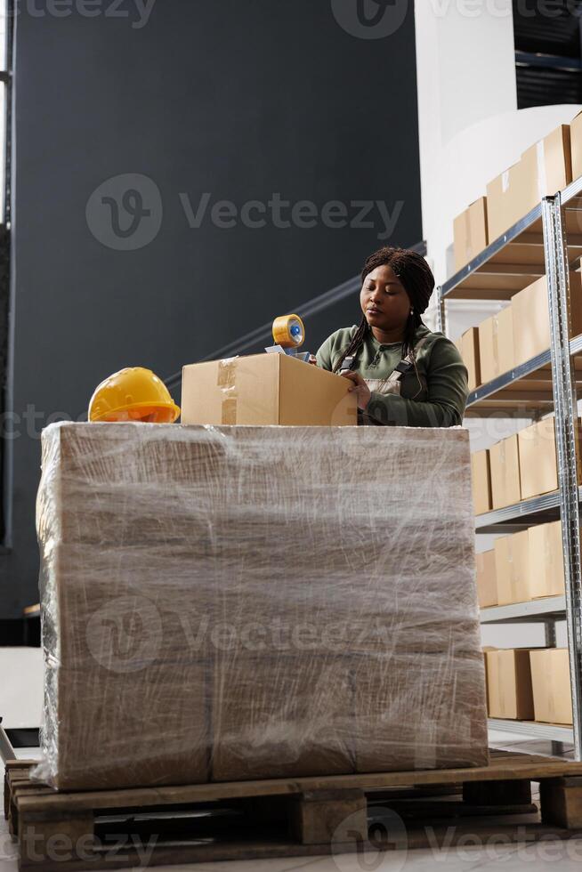 African american manager using adhesive tape to pack customers orders, preparing packages for delivery in storage room. Stockroom employee wearing industrial overall during merchandise inventory photo
