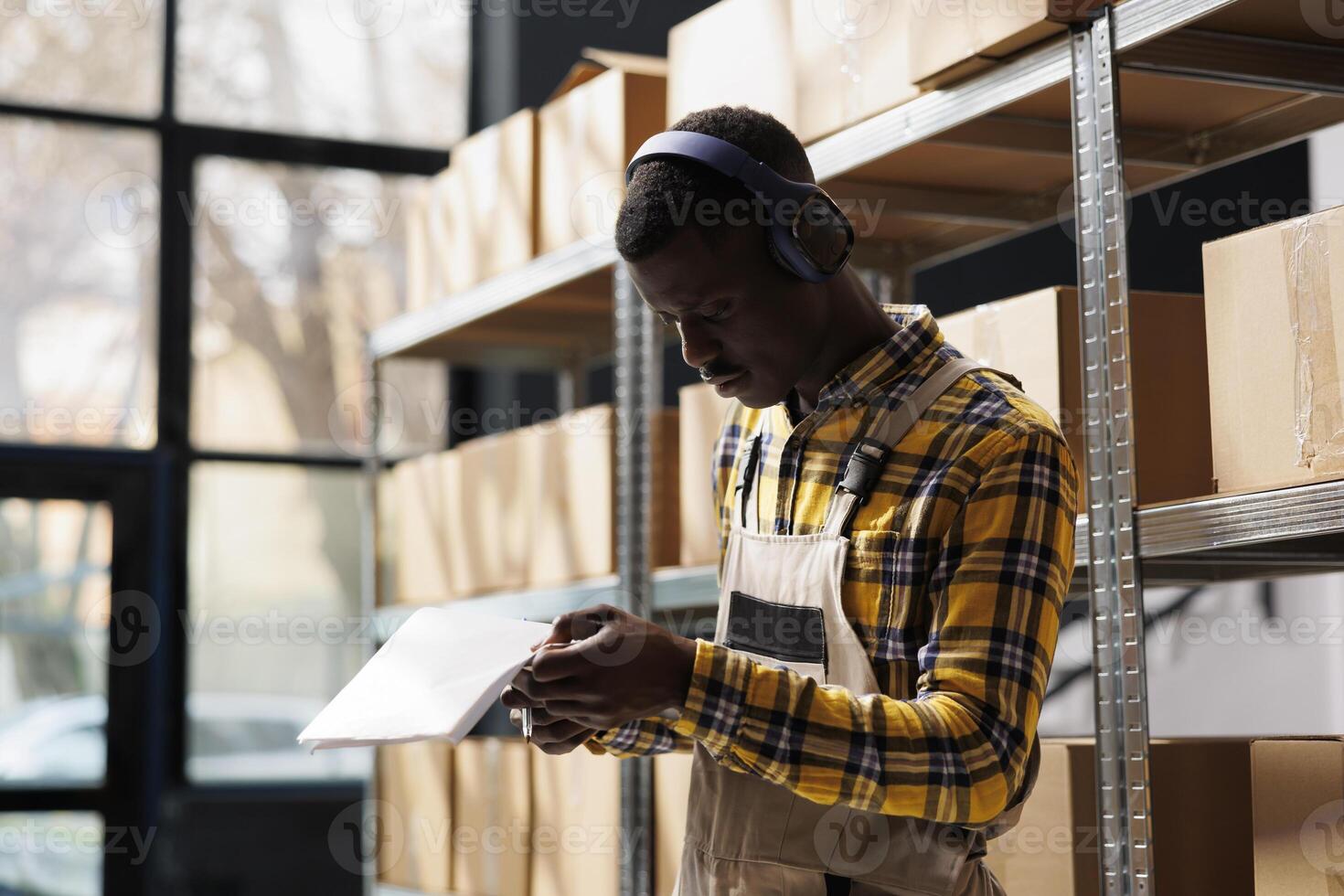 Shipment manager wearing headphones reading instruction documentation in warehouse. African american man listening to music, holding papers clipboard and checking goods logistics in storehouse photo